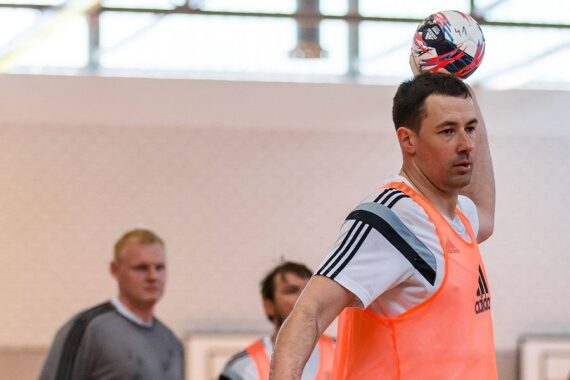 Ein Mann in einem weißen Hemd und einer orangefarbenen Weste bereitet sich darauf vor, während eines Hallentrainings einen Fußball über seinen Kopf zu werfen, während zwei andere Männer im Hintergrund verschwommen zu sehen sind.