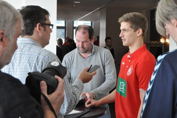 Ein junger Mann in einem roten Sporthemd wird von mehreren Journalisten interviewt; einer hält ein Aufnahmegerät in der Hand, ein anderer eine Kamera, und andere machen sich an einem kleinen Tisch im Haus Notizen.