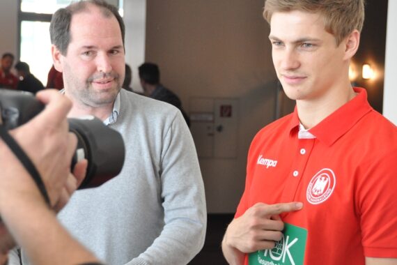 Ein junger Mann in einem roten Poloshirt mit einem Sportlogo zeigt auf seine Brust, während er für ein Foto posiert. Ein anderer Mann steht neben ihm, und eine Person im Vordergrund hält eine Kamera und fotografiert sie in einem Innenraum.