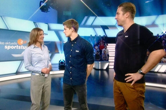 Eine Frau und zwei Männer stehen in einem hell erleuchteten Fernsehstudio, in dem das aktuelle sportstudio auf einem Bildschirm im Hintergrund zu sehen ist. Die Zuschauer sitzen hinter ihnen. Die drei scheinen ein Gespräch zu führen.