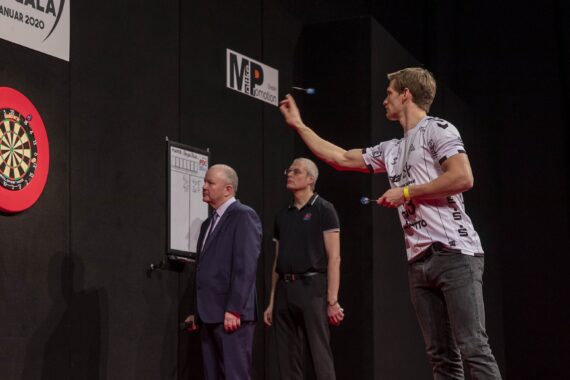 Ein Mann in einem weißen Hemd wirft einen Dartpfeil auf eine Dartscheibe, während zwei Beamte, einer in einem Anzug und einer in einem schwarzen Hemd, in einer professionellen Halle genau beobachten.