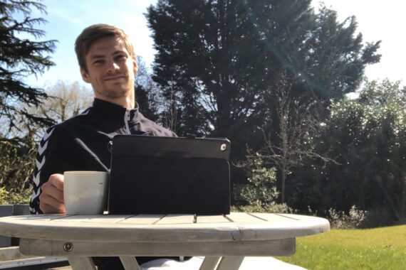 Ein Mann sitzt im Freien an einem runden Tisch und hält einen weißen Becher in der Hand. Vor ihm auf dem Tisch steht eine Tafel. Er lächelt, und im Hintergrund sind Bäume und helles Sonnenlicht zu sehen.