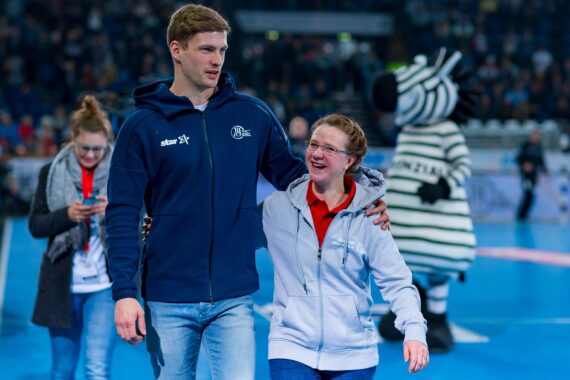 Ein Mann, der seinen Arm um eine lächelnde Frau gelegt hat, geht auf einem Sportplatz. Im Hintergrund sind eine Person in einem Zebra-Maskottchenkostüm und andere Personen zu sehen. Der Schauplatz scheint eine Sportveranstaltung in einer Halle zu sein.