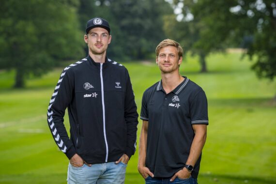 Zwei Männer stehen nebeneinander auf einem grasbewachsenen Feld mit Bäumen im Hintergrund. Beide tragen schwarze Sportkleidung mit weißen Logos; der eine trägt eine Mütze und eine Jacke, der andere ein kurzärmeliges Poloshirt.