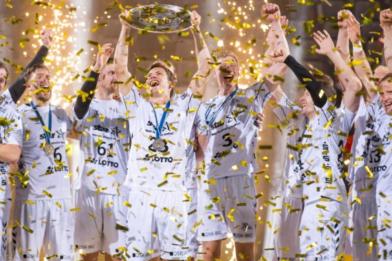 Ein jubelndes Handballteam in weißen Trikots steht auf der Bühne und hält inmitten von fliegendem Goldkonfetti und leuchtenden Funken einen Pokal hoch, mit Medaillen um den Hals.
