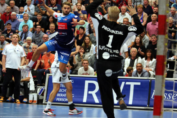 Ein Handballspieler in einem blauen Trikot springt, um den Ball in Richtung Tor zu werfen, während ein Torwart in einem schwarzen Trikot sich darauf vorbereitet, den Schuss zu blockieren. Die Zuschauer verfolgen das Geschehen aufmerksam von der Tribüne aus.