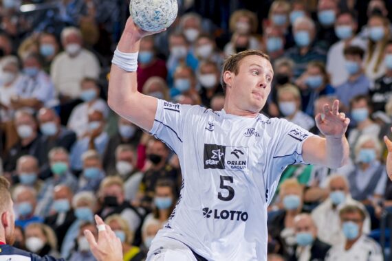 Ein männlicher Handballspieler in einem weißen Trikot bereitet sich mit dem Ball in der rechten Hand auf den Wurf vor, während ein dicht gedrängtes Publikum mit Gesichtsmasken das Spiel verfolgt.