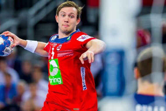 Ein männlicher Handballspieler in einem roten Trikot bereitet sich darauf vor, den Ball während eines Spiels zu werfen. Im Vordergrund ist ein anderer Spieler zu sehen, im Hintergrund ein unscharfes Publikum.