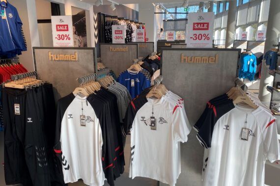 Hummel-Sporttrikots in Schwarz und Weiß sind in einem Geschäft auf Regalen ausgestellt, darüber befinden sich rote Verkaufsschilder mit einem Rabatt von 30 %. Auf der linken Seite sind blaue Hemden zu sehen, und das Sonnenlicht fällt durch große Fenster.