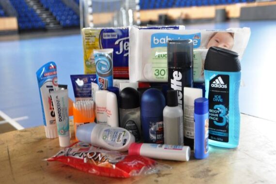 Auf einem Holztisch vor leeren blauen Stadionsitzen in einer Sporthalle sind verschiedene Körperpflegeprodukte wie Zahnpasta, Deodorant, Duschgel, Taschentücher und Babyfeuchttücher angeordnet.