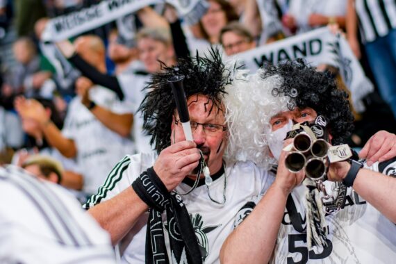 Zwei begeisterte Sportfans mit schwarz-weißen Lockenperücken und Gesichtsbemalung blasen in einer lebhaften Stadionmenge in Plastikhupen, tragen passende Trikots und sind von anderen mit Schals umgeben.