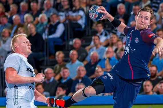 Zwei männliche Handballspieler in Aktion, von denen einer in der Luft springt, um den Ball ins Tor zu werfen. Eine große Menge von Zuschauern schaut im Hintergrund aufmerksam zu.