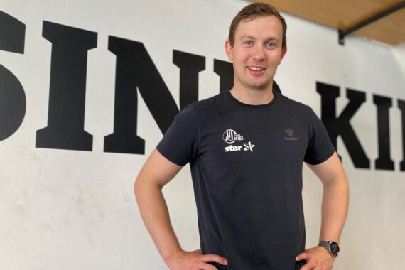Ein lächelnder junger Mann in einem dunklen T-Shirt steht in einem Innenraum, die Hände in die Hüften gestemmt, vor einer Wand mit großen schwarzen Buchstaben.