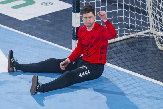 Ein männlicher Handballtorwart in einem roten Trikot und einer schwarzen Hose sitzt auf dem Boden vor dem Tor und hebt mit konzentrierter Miene die rechte Hand.