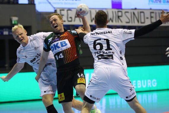 Ein männlicher Handballspieler in einem schwarz-orangenen Trikot springt mit dem Ball und versucht zu schießen, während zwei gegnerische Spieler in weißen Trikots versuchen, ihn während eines Hallenspiels zu blockieren.