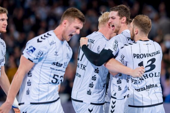 Eine Gruppe männlicher Handballspieler in weißen Trikots feiert leidenschaftlich auf dem Spielfeld, wobei sich zwei Spieler umarmen und andere jubeln, umgeben von einer verschwommenen Menge im Hintergrund.