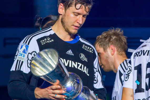 Ein männlicher Sportler in einem schwarz-weißen Trikot hält eine große silberne Trophäe und blickt darauf herab. Andere Mannschaftskameraden in ähnlichen Trikots stehen in der Nähe, im Hintergrund liegt Konfetti, das auf eine Siegesfeier hindeutet.