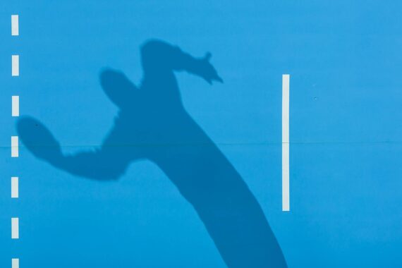 Schatten einer Person, die einen Ball hält, möglicherweise in der Mitte des Wurfs, auf einem hellblauen Sportplatz mit weißen Linien. Das Bild ist von oben aufgenommen, wodurch der längliche Schatten hervorgehoben wird.
