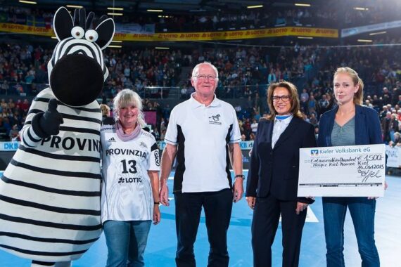 Fünf Personen stehen auf einem Sportplatz: ein Zebra-Maskottchen, zwei Frauen in Sporttrikots, ein Mann in einem weißen Polohemd und eine Frau im Anzug, die einen großen Scheck über 4 500 Euro in der Hand hält. Die Zuschauer füllen die Tribünen hinter ihnen.