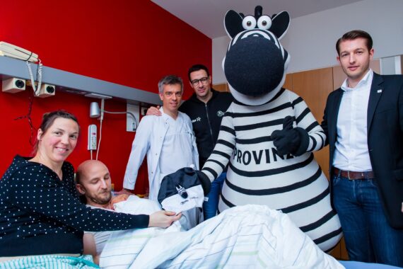 Ein Mann liegt in einem Krankenhausbett, umgeben von einer lächelnden Frau, zwei Männern, einem Arzt und einer Person in einem Zebra-Maskottchenkostüm. Die Frau überreicht dem Patienten ein Geschenk im Zebrakostüm. Das Zimmer hat rote und weiße Wände.