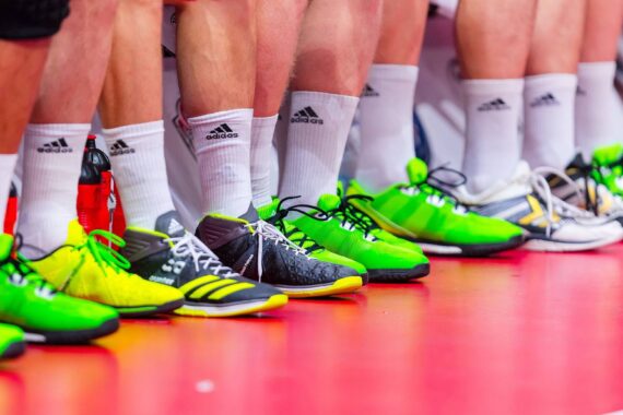 Eine Reihe von Menschen, die auf einem roten Boden stehen, mit weißen Adidas-Socken und bunten Sportschuhen, meist in neongrünen und schwarzen Farbtönen.