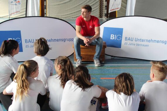 Ein Mann in einem roten Hemd sitzt auf einer Bank in einer Turnhalle und blickt auf eine Gruppe von Kindern in weißen Hemden, die auf dem Boden sitzen. Zwei Banner hinter ihm zeigen das Logo der BIG Unternehmensgruppe.