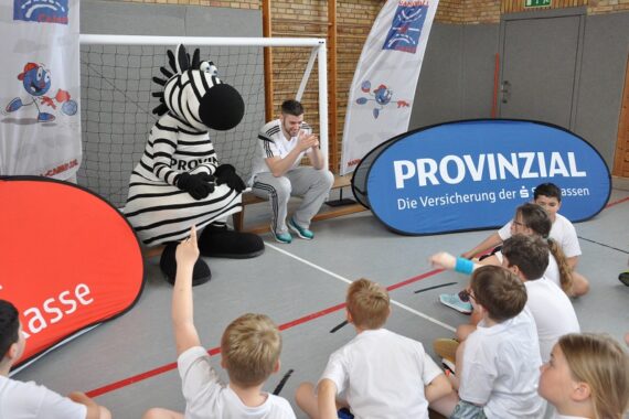 Eine Person und ein Zebramaskottchen sitzen neben einem Fußballtor in einer Turnhalle und sprechen mit einer Gruppe von Kindern in weißen Hemden, die auf dem Boden sitzen. Um sie herum sind Werbebanner in deutscher Sprache angebracht.