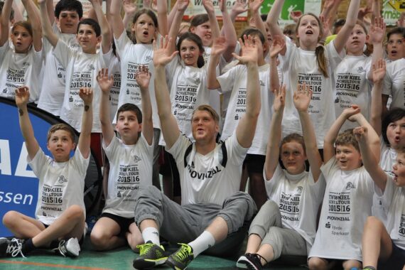 Eine Gruppe von Kindern und ein Erwachsener, alle in passenden weißen Sporthemden, sitzen und knien auf dem Boden einer Turnhalle, lächeln und heben begeistert die Hände in Richtung der Kamera.