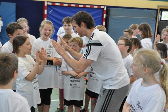Ein Mann in Sportkleidung gibt einer lächelnden Gruppe von Kindern in passenden Veranstaltungs-T-Shirts in einer Turnhalle enthusiastisch ein High-Five, während andere Kinder zusehen und mitmachen.