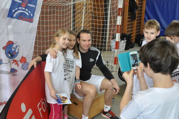 Eine Gruppe von Kindern in Sportkleidung posiert für ein Foto mit einem erwachsenen Trainer, der auf einer Bank in einer Turnhalle sitzt, während ein anderes Kind ein Foto mit einem Tablet macht. Im Hintergrund sind ein Netz und eine bunte Fahne zu sehen.