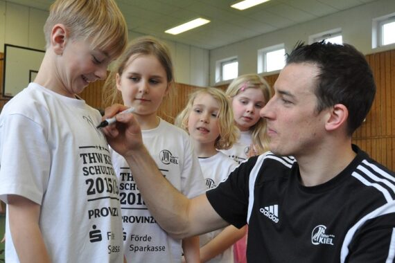 Ein Mann in einem schwarzen Sporthemd signiert das weiße T-Shirt eines lächelnden Jungen, während drei andere Kinder in einer Sporthalle zusehen. Alle wirken engagiert und glücklich.