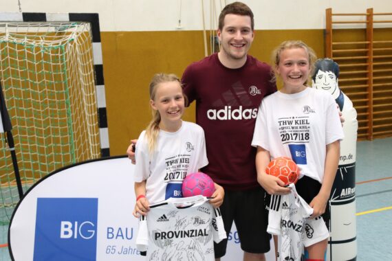 Drei Personen posieren in einer Turnhalle: Ein Erwachsener steht hinter zwei lächelnden Mädchen, die signierte Sporttrikots und bunte Handbälle halten. Im Hintergrund sind eine aus Pappe ausgeschnittene Figur und Sportgeräte zu sehen.