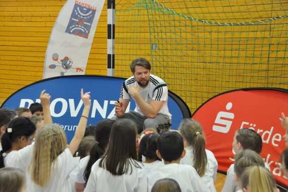 Ein Trainer hockt vor einer Gruppe junger Kinder, die während einer Sportveranstaltung in einer Turnhalle eifrig die Hände heben. Im Hintergrund sind Banner und ein Netz zu sehen.