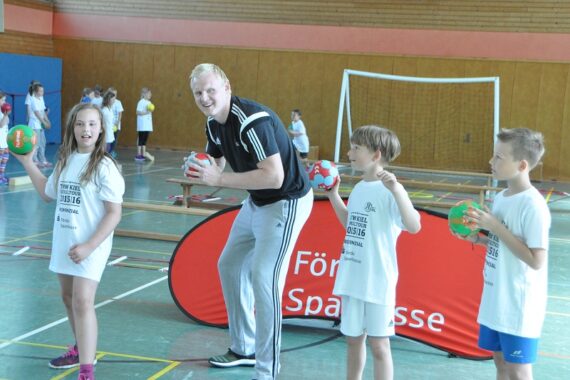 Ein lächelnder Trainer und vier Kinder in Sportkleidung stehen in einer Turnhalle und halten Handbälle. Hinter ihnen stehen ein Tor und eine rote Fahne auf dem Boden. Die Kinder scheinen spielbereit zu sein, und alle sehen glücklich und energiegeladen aus.