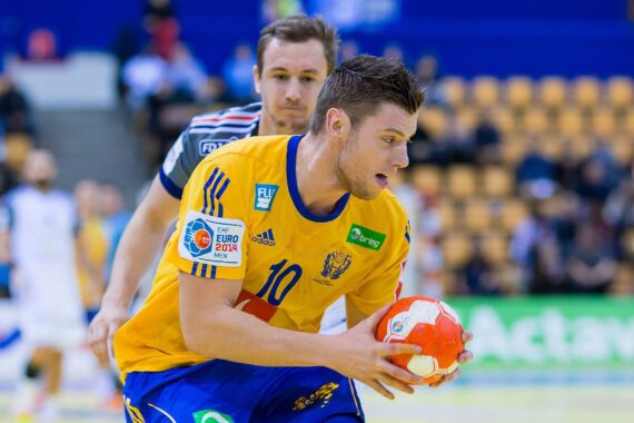 Ein männlicher Handballspieler in einem gelben Trikot hält einen orange-weißen Ball, dicht gefolgt von einem Gegner in einem gestreiften Trikot, während eines Spiels in einer Halle mit Zuschauern im Hintergrund.