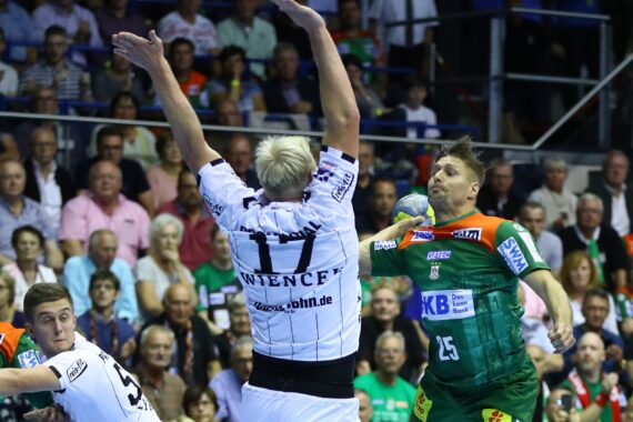 Ein Handballspieler in einem grünen Trikot springt, um den Ball zu werfen, während ein Verteidiger in einem weißen Trikot die Arme zum Abwehren hebt. Die Aktion findet vor einer vollen Zuschauermenge in einer Indoor-Arena statt.