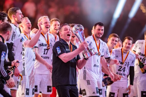 Eine jubelnde Männerhandballmannschaft in weißen Trikots feiert auf einer hell erleuchteten Bühne, während ein Mann in Schwarz einen großen Pokal hochhält. Die Mannschaftsmitglieder lächeln, jubeln und tragen Medaillen um den Hals.