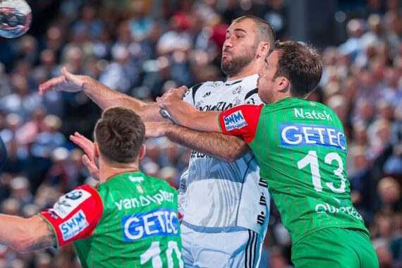 Drei männliche Handballspieler sind während eines Spiels in Aktion. Ein Spieler in einem weißen Trikot wird von zwei Spielern in grünen Trikots verteidigt, auf denen GETEC und 13 zu sehen sind. Die Szene ist intensiv mit einer unscharfen Menschenmenge im Hintergrund.