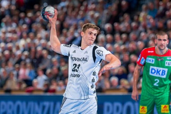 Ein männlicher Handballspieler in einem weißen Trikot bereitet sich darauf vor, den Ball während eines Spiels zu werfen, während ein anderer Spieler in einem grün-roten Trikot im Hintergrund steht und die Zuschauer zusehen.