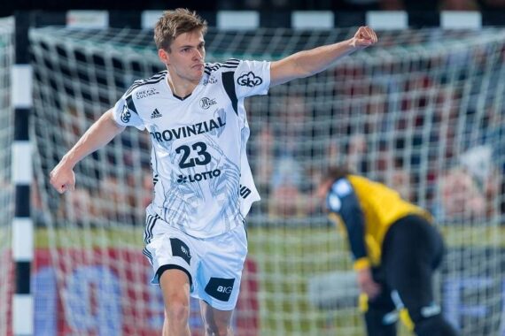 Ein männlicher Handballspieler, der ein weißes Trikot mit der Nummer 23 trägt, jubelt während eines Spiels mit erhobener Faust; im Hintergrund ist ein gelb-schwarz gekleideter Torwart in der Nähe des Tors zu sehen.