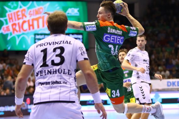 Ein Handballspieler in einem grün-orangenen Trikot springt, um den Ball zu werfen, während ein Spieler in einem weißen Trikot mit der Nummer 13 verteidigt. Im Hintergrund sind eine Menschenmenge und ein großes Schild in deutscher Sprache zu sehen.