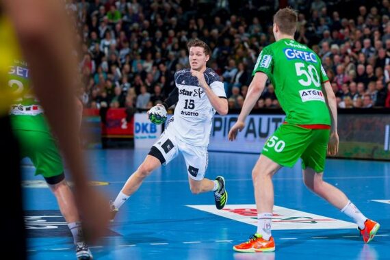 Ein männlicher Handballspieler in einem weißen Trikot mit der Nummer 15 läuft mit dem Ball, während ein Abwehrspieler in einem grünen Trikot mit der Nummer 50 ihn blockiert. Die Szene spielt sich auf einem Hallenplatz mit einer Menschenmenge im Hintergrund ab.