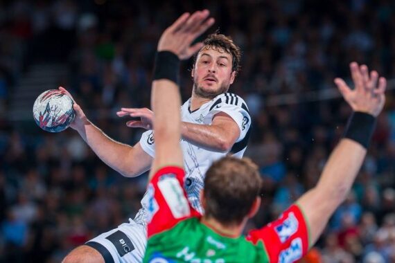 Ein Handballspieler in einem weißen Trikot springt und bereitet sich auf den Wurf vor, während ein anderer Spieler in einem rot-grünen Trikot die Arme hebt, um während eines intensiven Spiels zu blocken. Die Zuschauer sind im Hintergrund verschwommen.