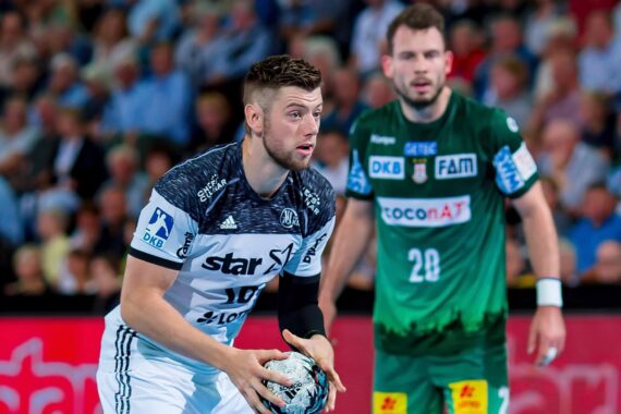 Ein männlicher Handballspieler in einem weiß-schwarzen Trikot hält den Ball, konzentriert und bereit zum Pass, während ein anderer Spieler in einem grünen Trikot hinter ihm steht. Eine Menschenmenge füllt den verschwommenen Hintergrund.