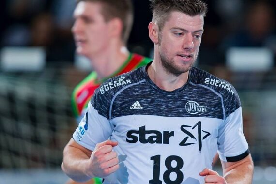 Ein männlicher Handballspieler in einem weiß-schwarzen Trikot mit der Nummer 18 und Sponsorenlogos befindet sich auf dem Spielfeld, während ein anderer Spieler unscharf im Hintergrund zu sehen ist. Die Szene erscheint während eines Spiels.
