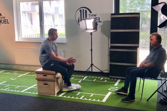 Zwei Männer sitzen sich für ein Interview auf einem Kunstrasen, der wie ein Sportplatz markiert ist, gegenüber, Licht und Kameratechnik sind aufgebaut. An den Wänden prangt das Logo des THW Kiel, Fenster lassen Tageslicht herein.