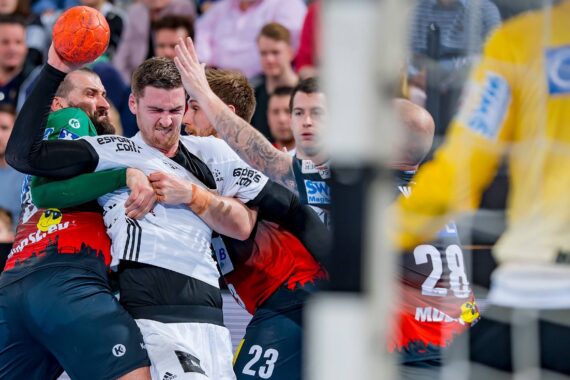 Mehrere männliche Handballspieler in Aktion während eines Spiels, die eng um den Ball kämpfen. Ein Spieler in Weiß wird von Gegnern in Rot und Schwarz geblockt. Im Vordergrund ist ein Torwart in Gelb zu sehen. Die Zuschauer im Hintergrund schauen zu.