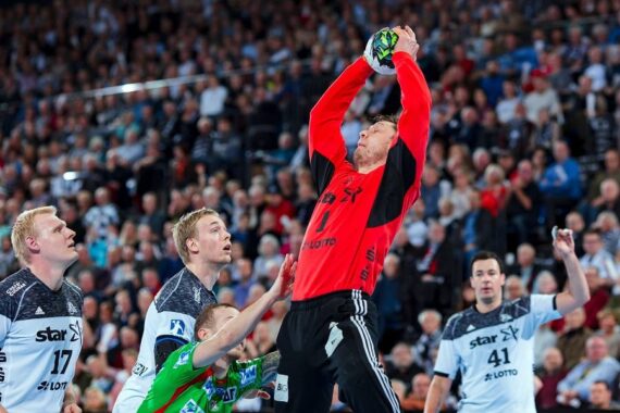 Ein Handballtorwart in einem roten Trikot springt, um den Ball über seinem Kopf zu fangen, umgeben von gegnerischen Spielern in weißen Trikots während eines Spiels, mit einem großen Publikum im Hintergrund.