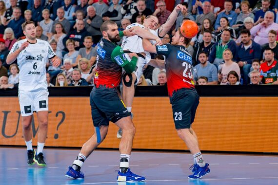 Zwei Handballspieler in dunklen Trikots blockieren einen gegnerischen Spieler in Weiß, als dieser zum Wurf ansetzt, während die Zuschauer im Hintergrund das intensive Geschehen auf dem Spielfeld verfolgen.