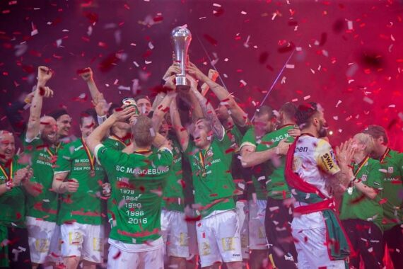 Eine jubelnde Sportmannschaft in grünen Trikots feiert auf der Bühne und hebt gemeinsam einen Pokal inmitten von fallendem roten Konfetti und dramatischer Beleuchtung. Die Spieler lächeln und jubeln und zeigen sichtbare Begeisterung und Stolz.